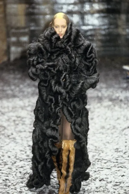 A runway model walking the Alexander McQueen Eshu Autumn/Winter 2000 show wearing an oversized full-length black coat made entirely of dense, swirling, hair-like curls, paired with knee-high textured tan boots.