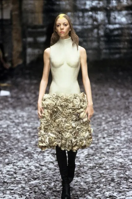 A model presenting a sculptural cream-colored dress from Alexander McQueen's Eshu Autumn/Winter 2000 collection, featuring a fitted bodice with anatomical bust detailing and a textured, rosette-style mini skirt paired with black boots.