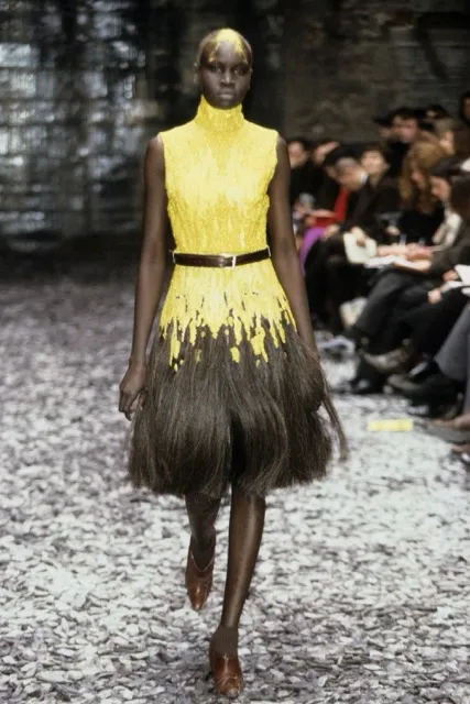 Model Alek Wek walking the runway for Alexander McQueen's Eshu Autumn/Winter 2000 collection in a sleeveless high-neck dress featuring a bright yellow beaded bodice that bleeds into a textured dark brown fringed skirt, accented with a slim dark waist belt.