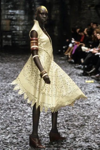 Model Alek Wek presenting a look on the runway for Alexander McQueen's Eshu Autumn/Winter 2000 collection, wearing an asymmetrical light yellow laser-cut leather dress with jagged hems, styled with stacked wooden bangles and brown heels.