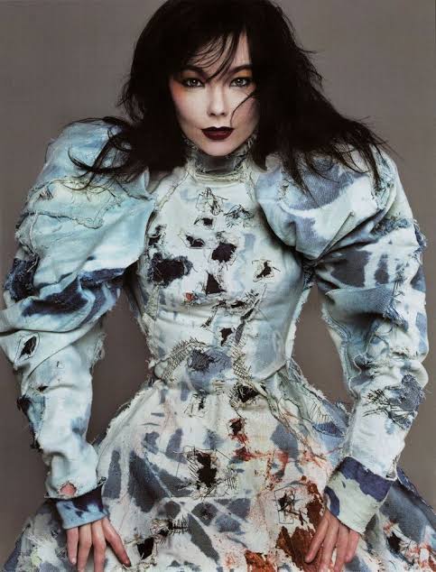 Studio portrait of musician Björk wearing an avant-garde distress-patterned high-neck dress with dramatic puffed shoulders from Alexander McQueen's Eshu collection. She looks forward with dark lip makeup and windswept dark hair.