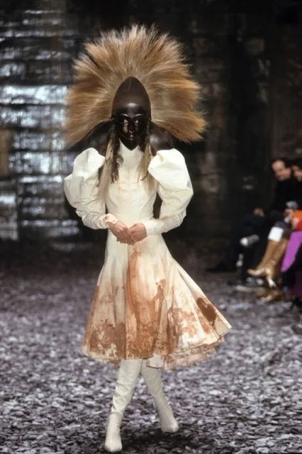 A runway model walking the Alexander McQueen Eshu Autumn/Winter 2000 show in a cream-colored high-neck dress with puffed sleeves and earthy mud-toned staining along the skirt, completed with an ornate carved African-style wooden mask topped with a large fan of hair-like fibers.