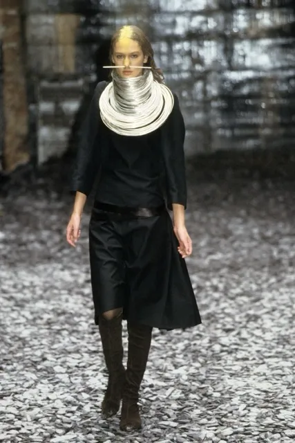 A model walking the runway for Alexander McQueen's Eshu Autumn/Winter 2000 collection, styled with gold pigments across her forehead and wearing a black outfit framed by a massive, layered metallic silver coil neck piece and facial pin.