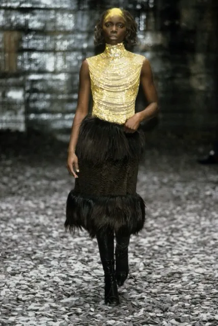 A runway look from Alexander McQueen's Eshu Autumn/Winter 2000 collection featuring a model in a sleeveless gold beaded top paired with a tiered black faux-fur knee-length skirt and black knee-high boots.