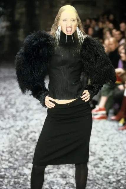 A runway model presenting a look from Alexander McQueen's Eshu Autumn/Winter 2000 show, wearing a black structured top with voluminous textured black faux-fur shoulders, a black pencil skirt, and a silver metal mouth-hook accessory extending from her teeth.